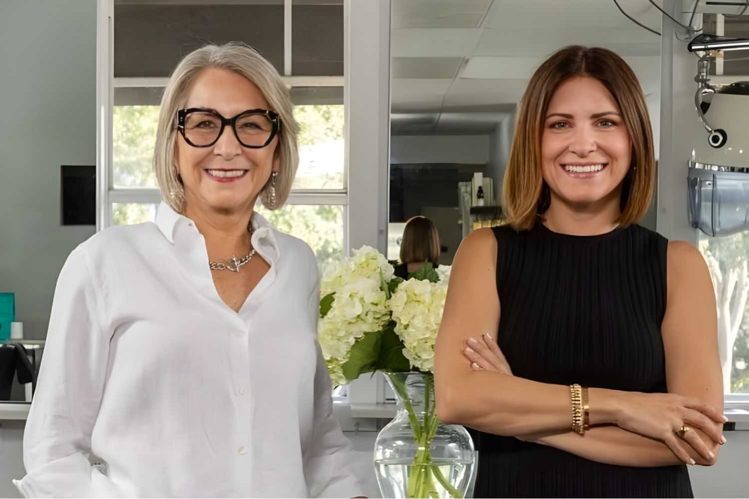 Two women smiling, standing beside a vase of white flowers in an indoor setting.